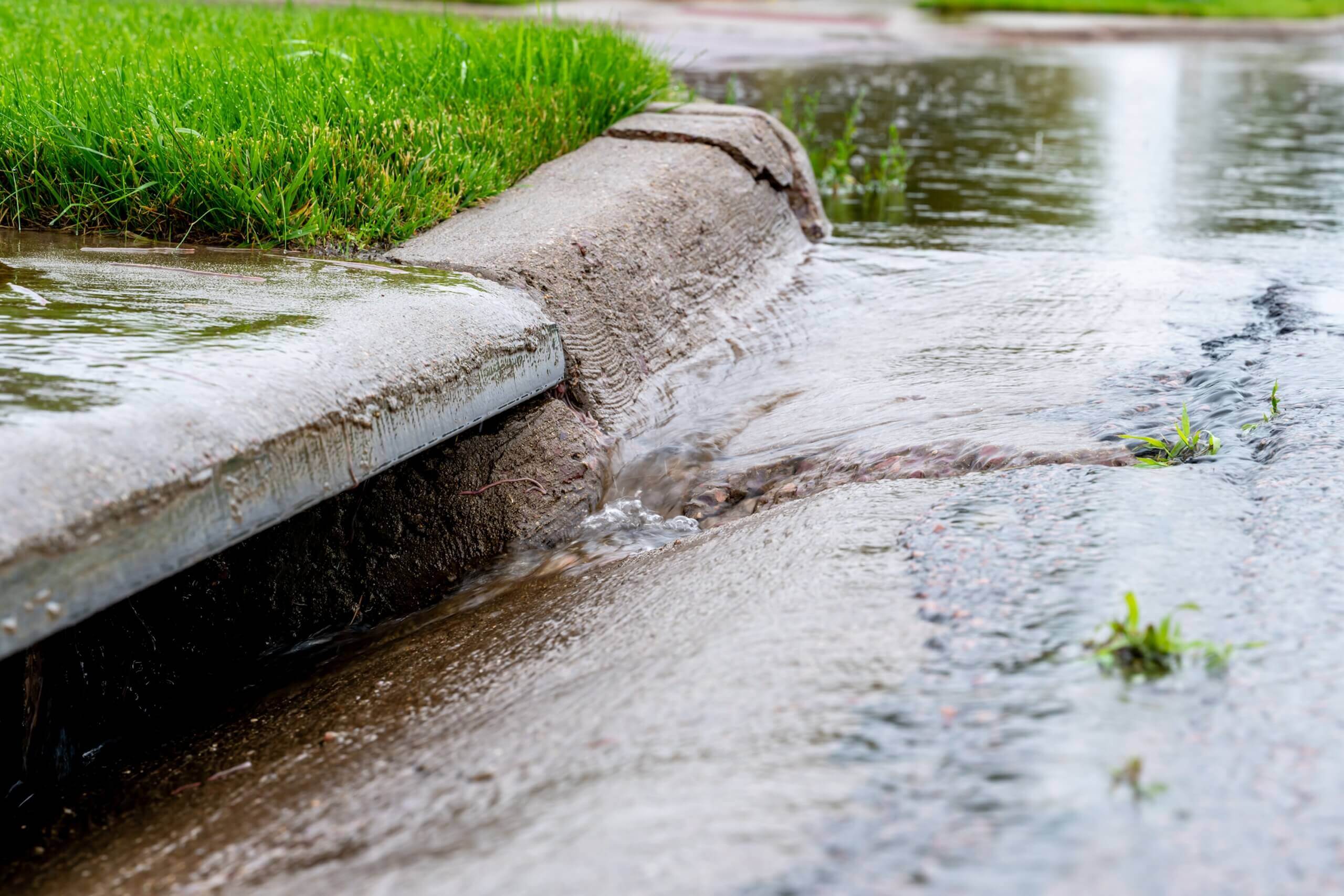 Regendurchflossener Bürgersteig mit abfließendem Wasser entlang der Kante; rasch wachsendes Gras im Vordergrund.