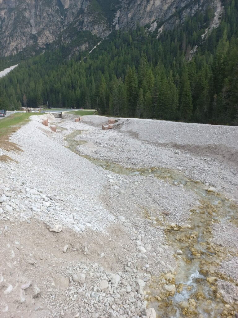 Berglandschaft mit einem kleinen Bach, umgeben von steinigem Ufer und dichten Nadelwäldern unter majestätischen Gipfeln.