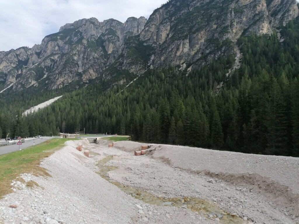 Berglandschaft mit Wald und Straße, Kiesbett im Vordergrund, unter bewölktem Himmel. Perfekt für Natur- und Reisefotografie.