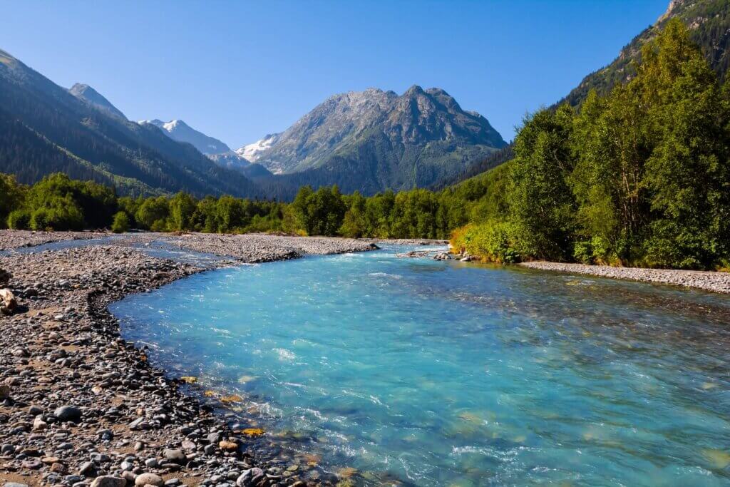Bergfluss mit klarem Wasser, Kieselstrand und bewaldeten Bergen unter blauem Himmel bei Sonnenschein.