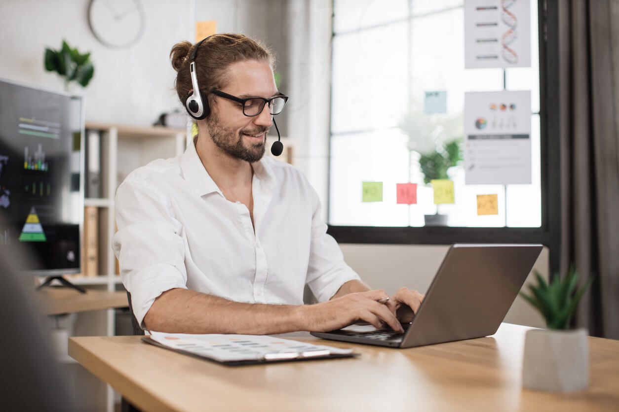 Mann im Büro mit Headset arbeitet am Laptop, lächelt, während er online kommuniziert. Homeoffice-Arbeitsumgebung.