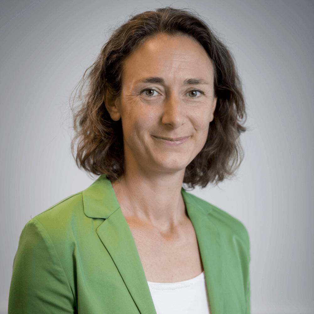 Porträt einer Frau in einem grünen Blazer mit neutralem Hintergrund, lächelnd in die Kamera blickend.